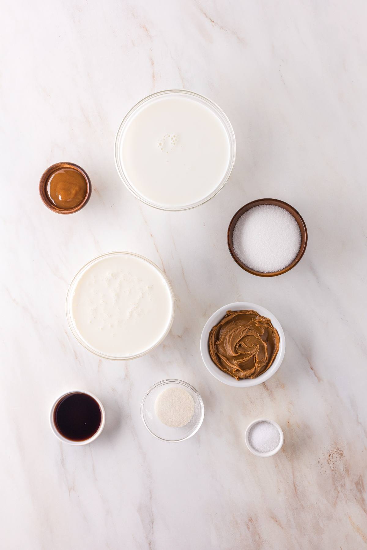 Seven bowls with milk, cream, sugar, peanut butter, vanilla, gelatin, and salt on a white marble surface.