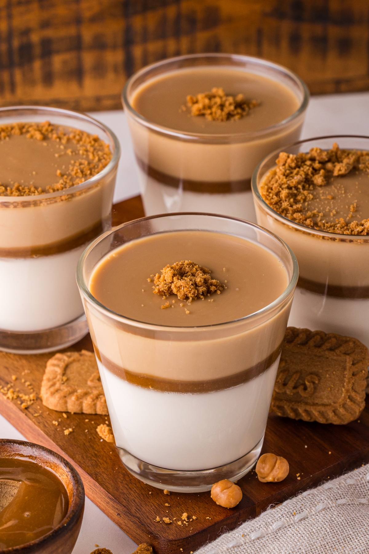 Four glasses of layered biscoff panna cotta dessert with caramel and crumbled cookies on top, placed on a wooden board.