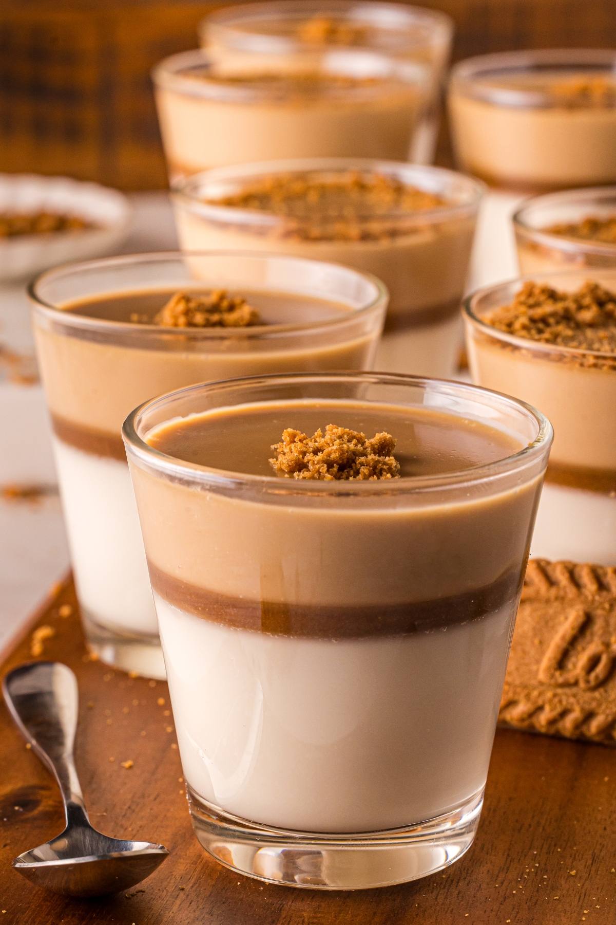 Glasses of layered biscoff panna cotta dessert with creamy white and brown layers, topped with cookie crumbs on a wooden board.