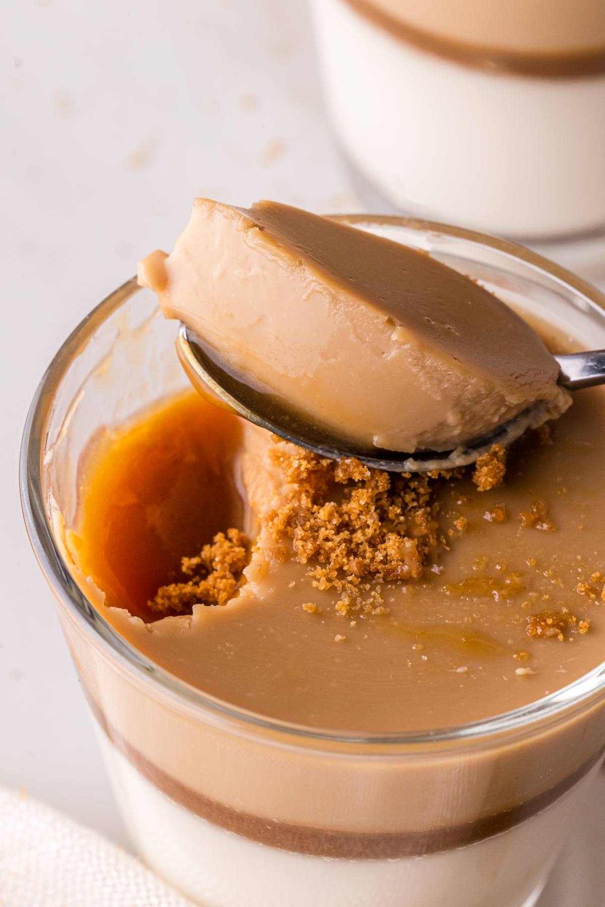 A spoon holds a bite of caramel pudding above a glass topped with brown sugar crumbs.