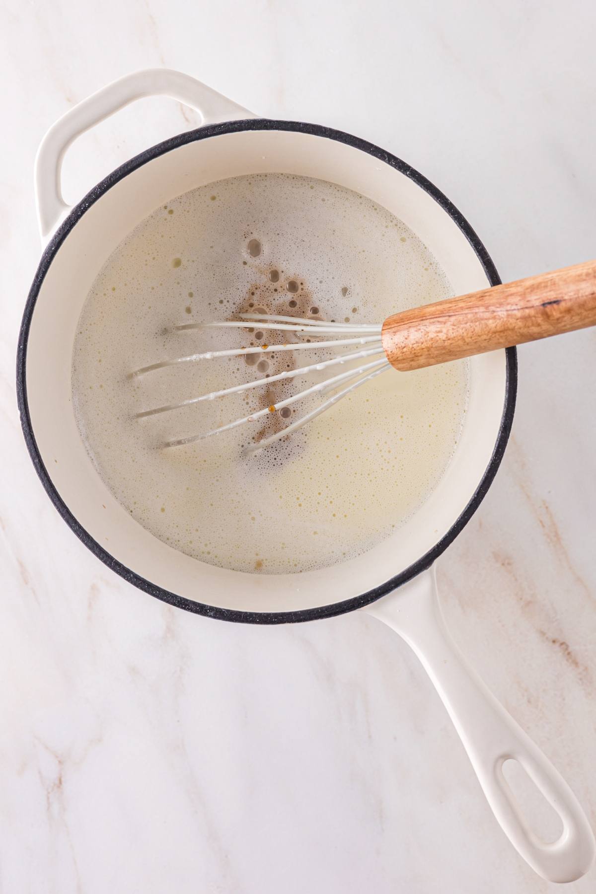 A whisk stirring a foamy liquid in a white pot on a marble countertop.