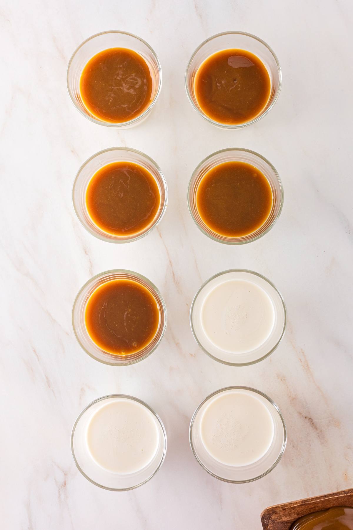 Eight small glass bowls arranged in two columns, each with caramel or light cream liquid, on a marble surface.