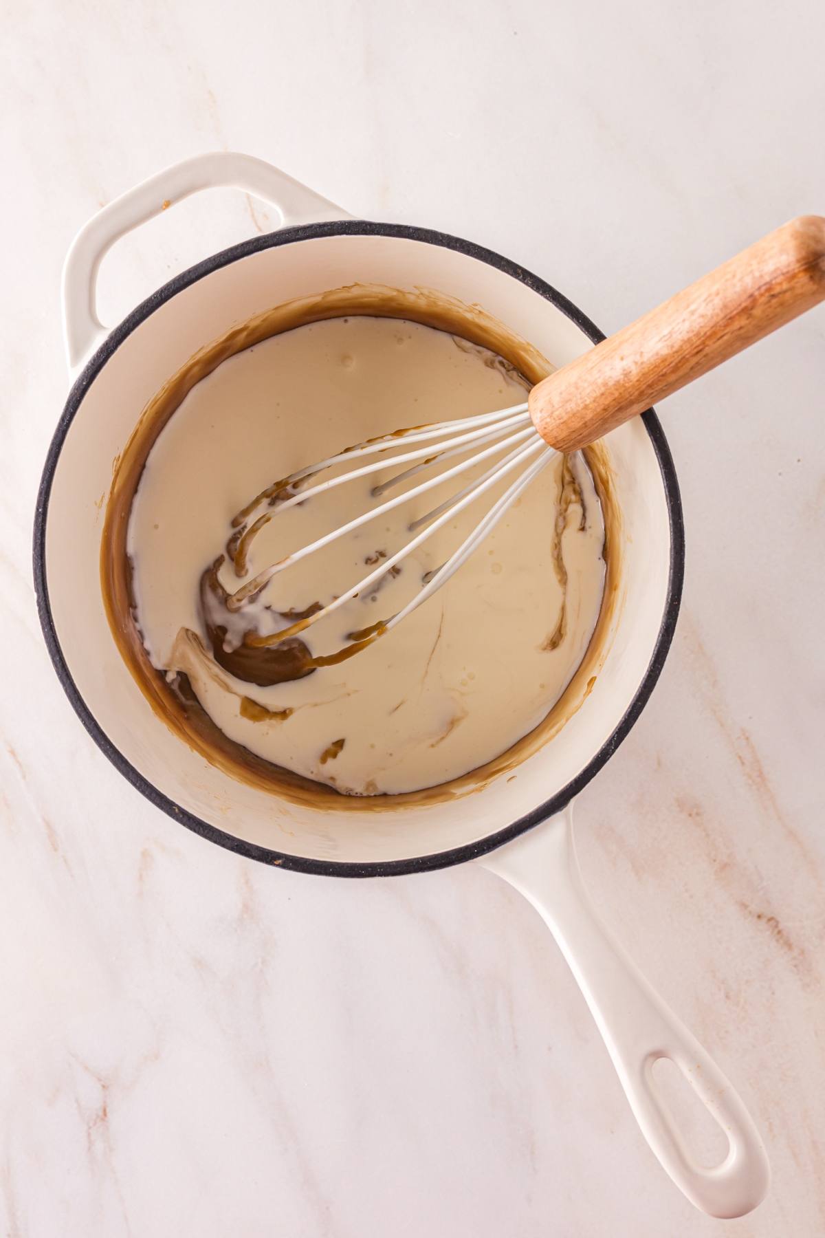 A whisk stirring light brown sauce in a white pot on a marble countertop.