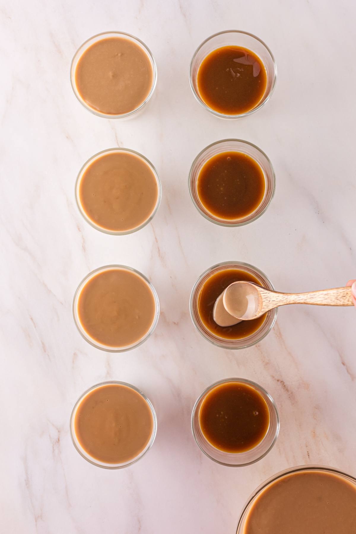 Eight small glass bowls with brown sauces, one being stirred with a wooden spoon, on a marble surface.