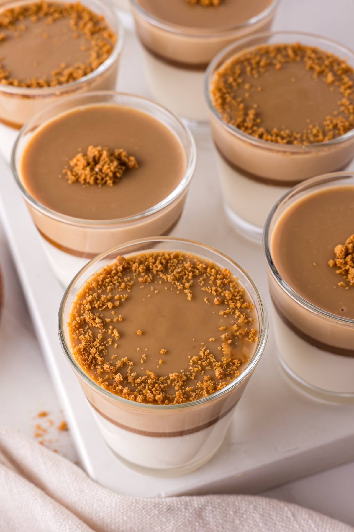 Glasses of layered panna cotta topped with caramel sauce and crumbled cookies on a white tray.