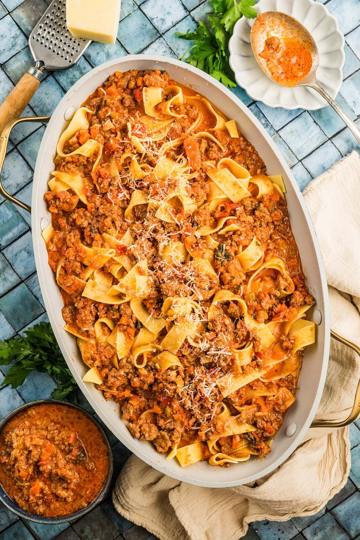 Wide oval dish of pappardelle pasta with authentic Bolognese sauce, topped with grated cheese, on a blue tiled surface.