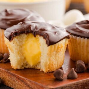 Boston cream cupcakes with chocolate frosting and yellow cream filling, one with a bite taken out, on a wooden board.
