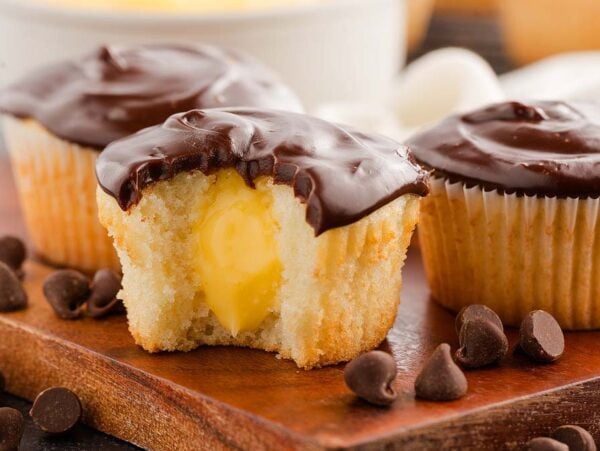 Boston cream cupcakes with chocolate frosting and yellow cream filling, one with a bite taken out, on a wooden board.