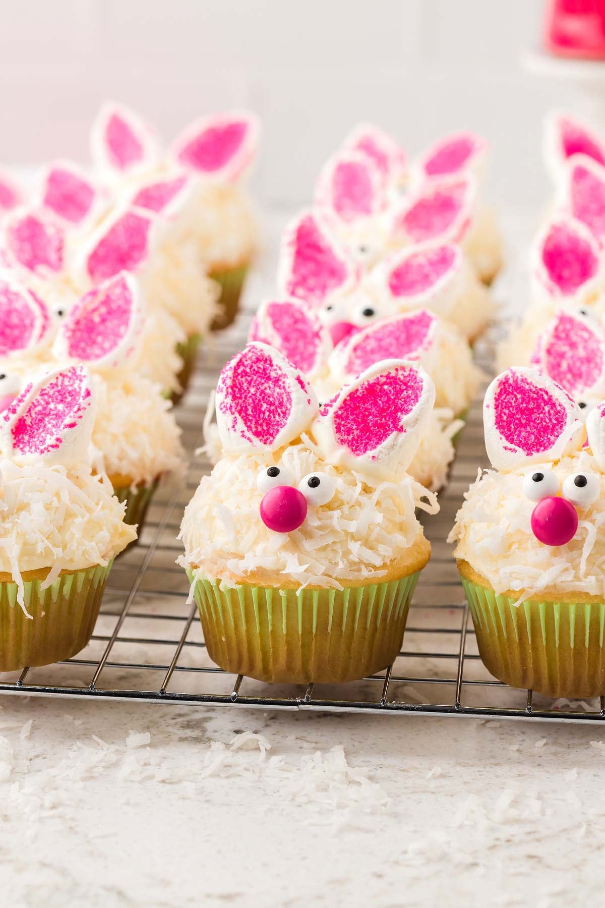 Bunny cupcakes decorated with pink ears, candy eyes, and noses are arranged on a cooling rack.