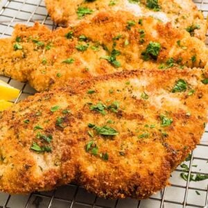 Golden breaded chicken cutlets rest on a cooling rack, garnished with fresh parsley and served alongside zesty lemon wedges.