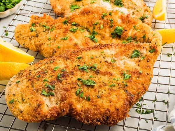 Golden breaded chicken cutlets rest on a cooling rack, garnished with fresh parsley and served alongside zesty lemon wedges.
