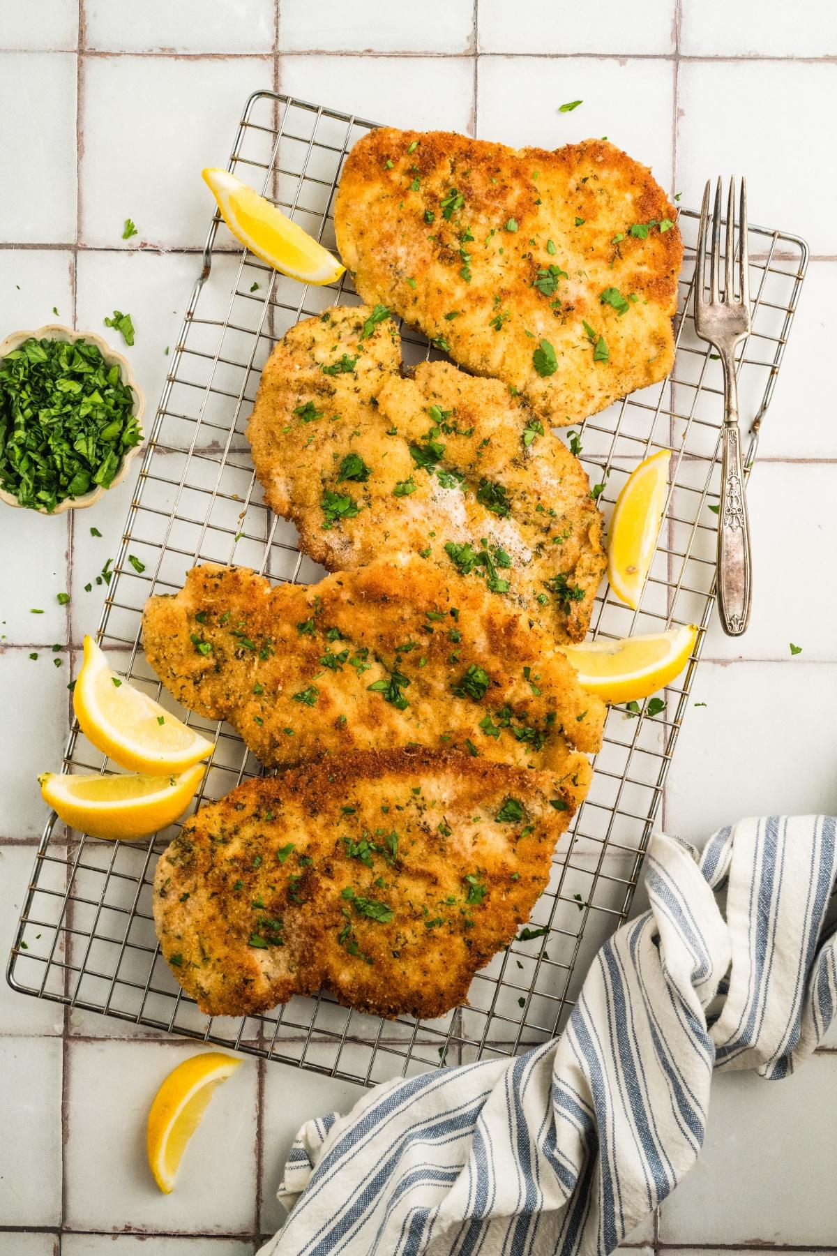 Four breaded chicken cutlets on a cooling rack, garnished with parsley and lemon wedges, with a fork nearby.