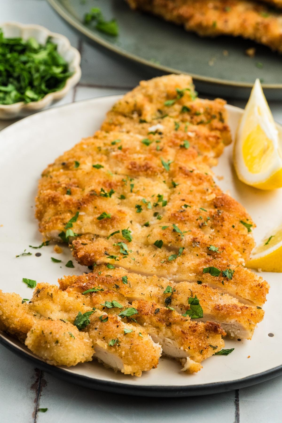 Breaded chicken cutlet garnished with chopped parsley, served with lemon wedges on a white plate.