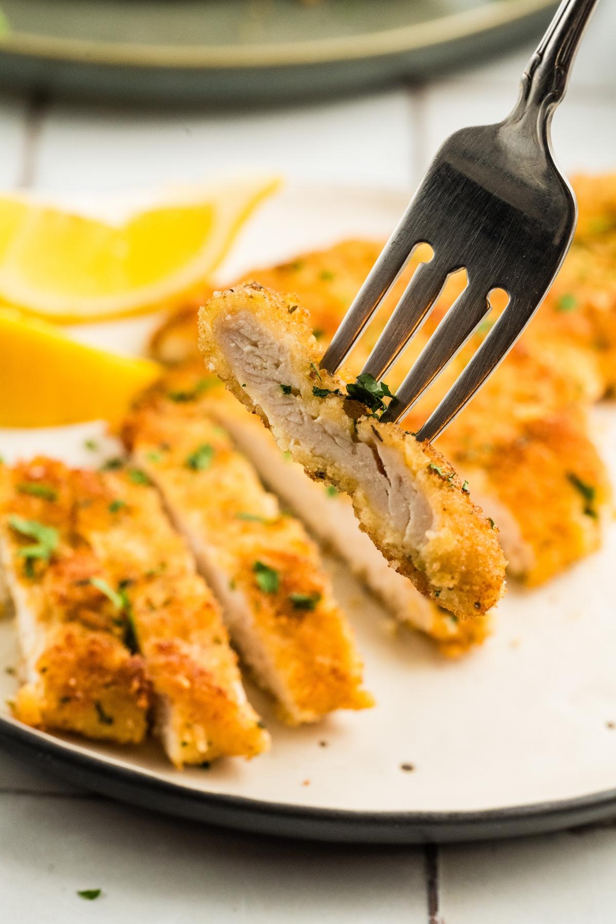 A fork holds a slice of breaded chicken cutlet, with more slices and lemon wedges on a plate below.