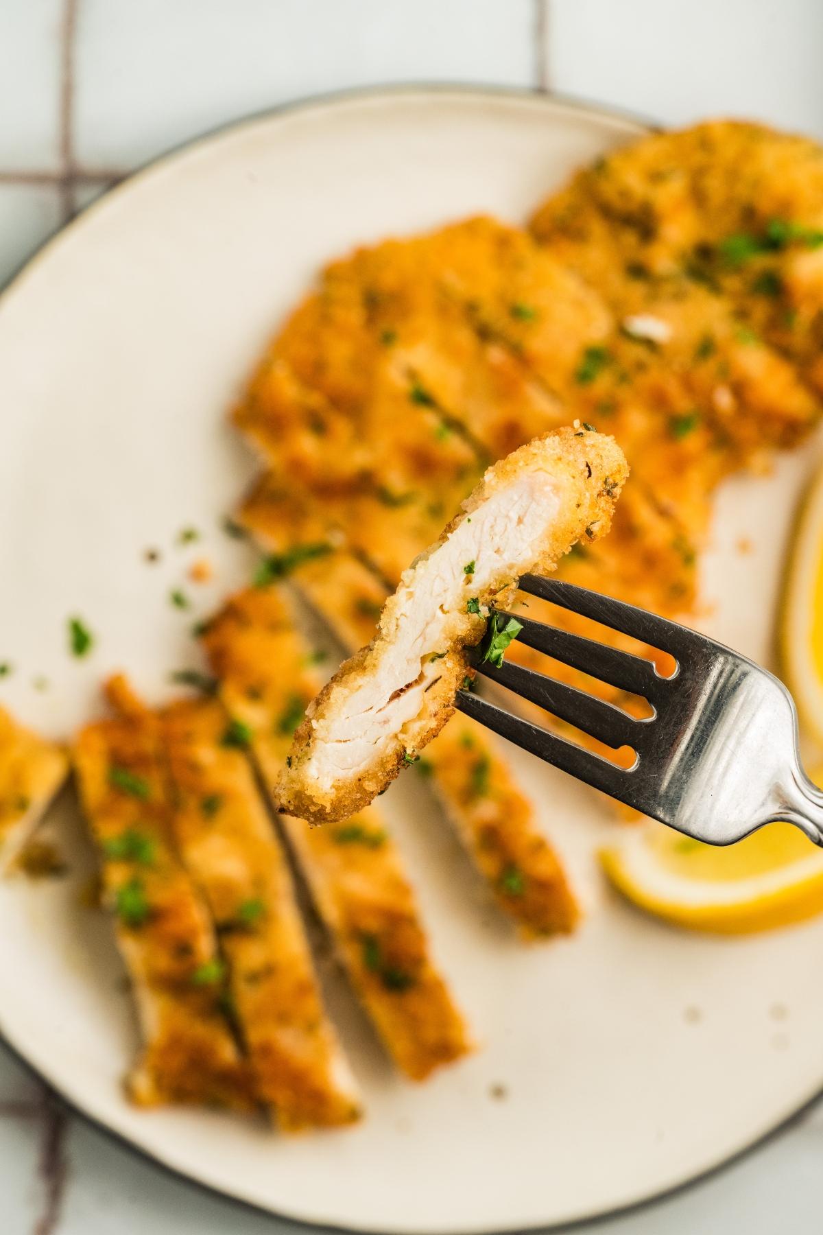 A fork holds a slice of breaded chicken cutlet above a plate with more cutlet slices and lemon wedges.