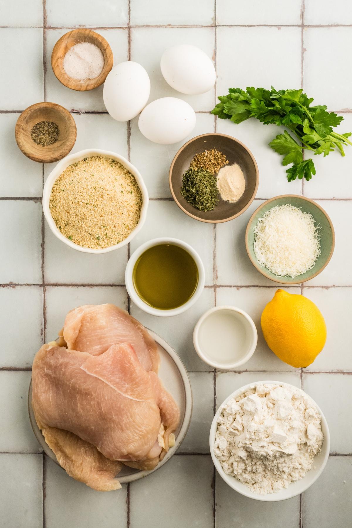 Raw chicken breasts, eggs, flour, breadcrumbs, seasonings, parsley, oil, grated cheese, and a lemon on a tiled surface.