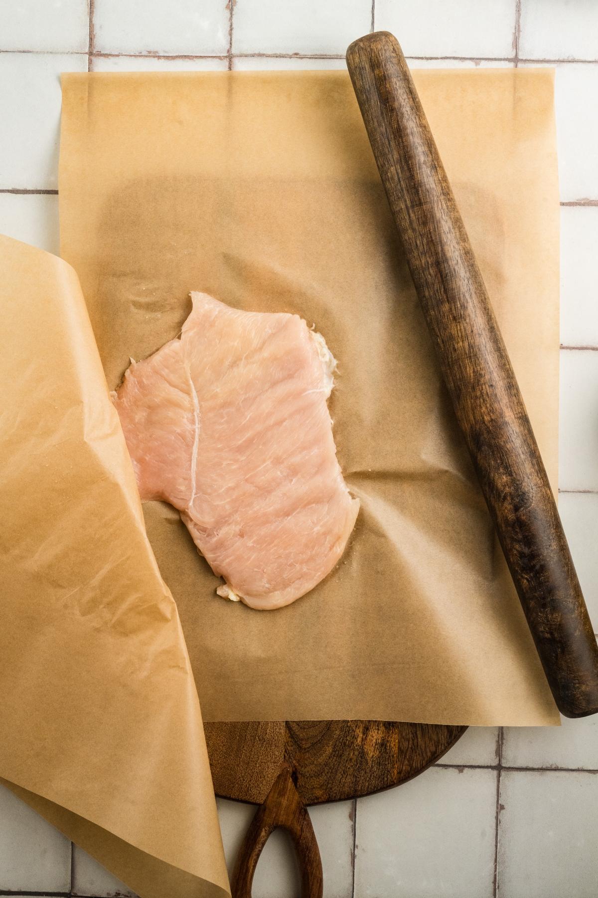 Raw chicken breast between parchment paper, flattened with a wooden rolling pin on a cutting board.