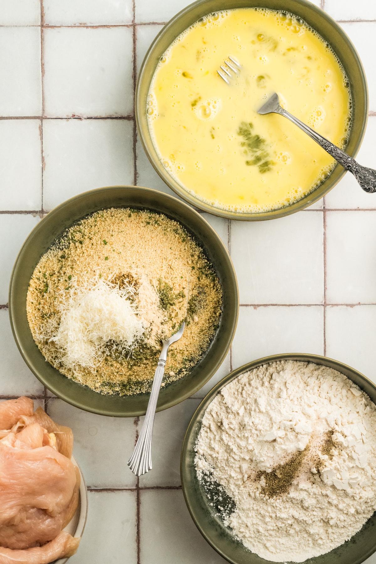 Three bowls with flour, egg mixture, and breadcrumbs, plus a plate of raw chicken on a tiled countertop.
