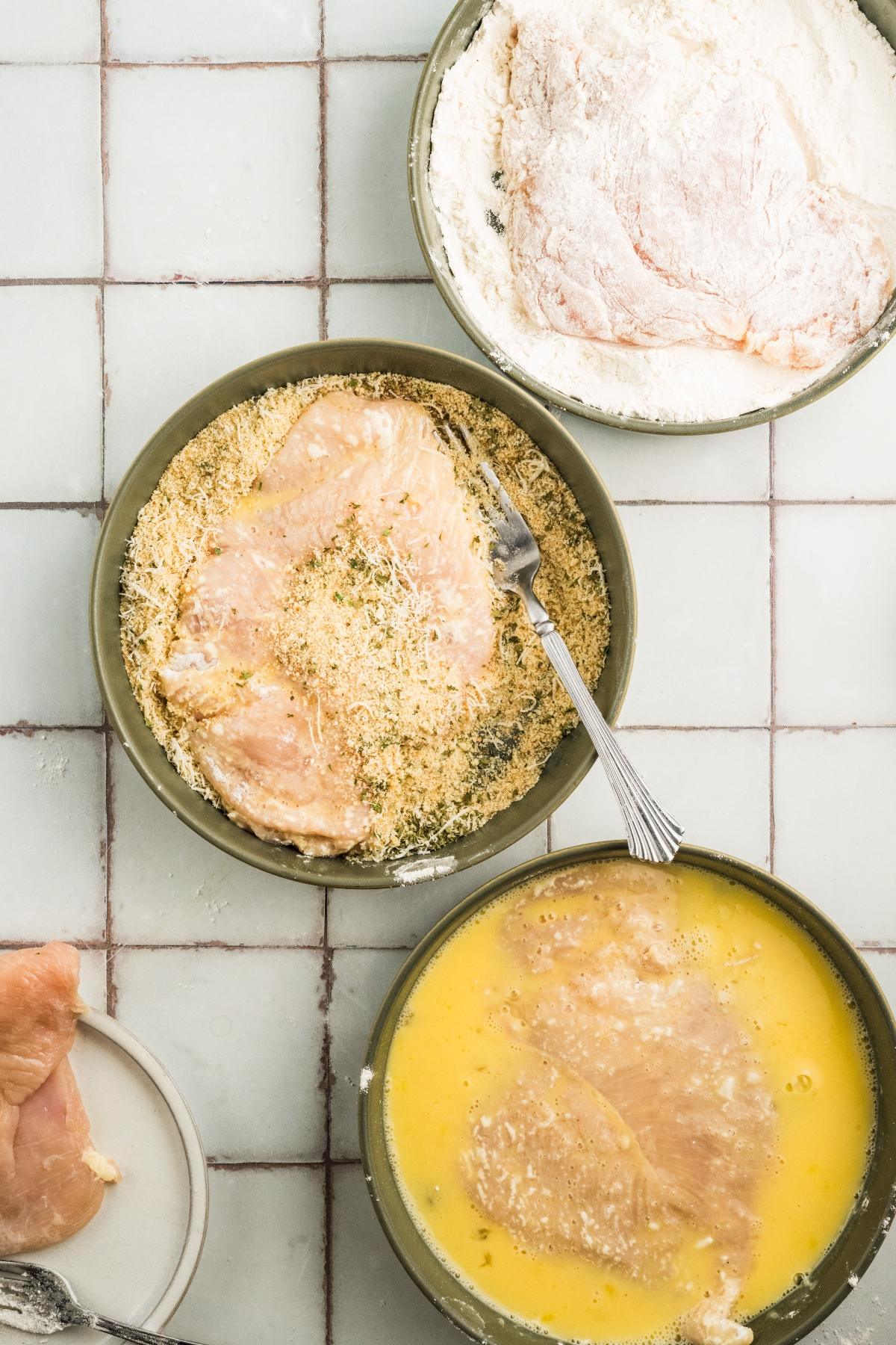 Three bowls with chicken: one in flour, one in egg wash, and one in breadcrumbs, on a tiled surface.