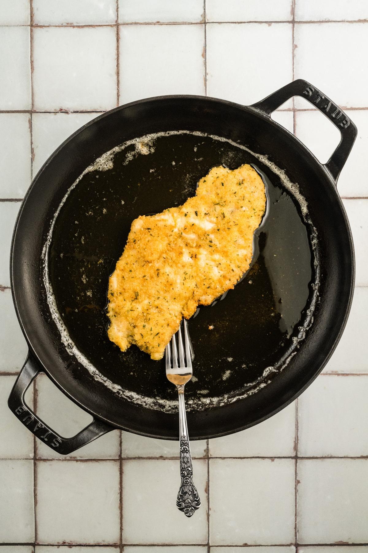 A breaded cutlet frying in oil in a black skillet, with a fork resting on the cutlet.