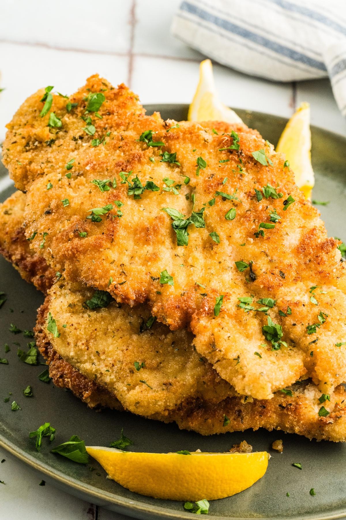Golden breaded chicken cutlets topped with chopped parsley on a plate with lemon wedges.