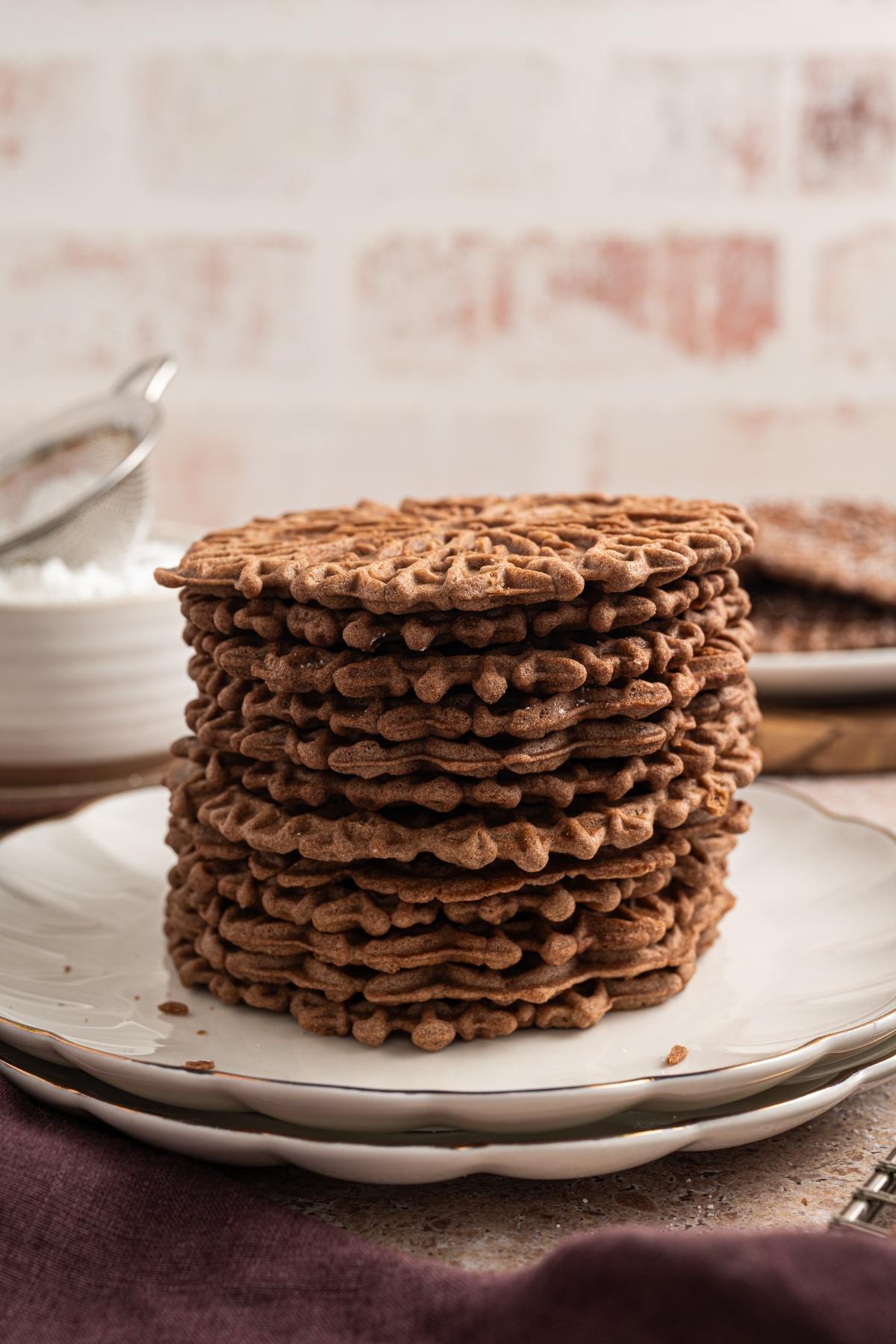 A stack of chocolate pizzelle cookies sits on a white plate with a strainer in the background.