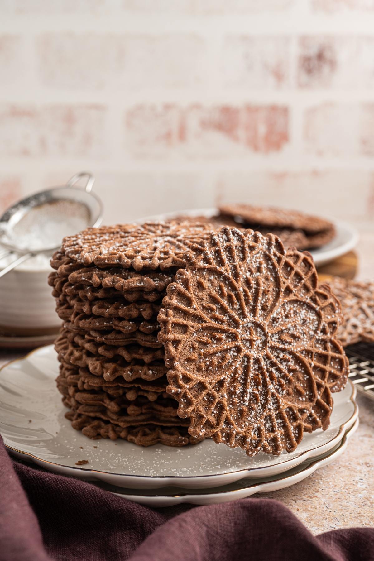 A stack of chocolate pizzelle cookies dusted with powdered sugar on a white plate.