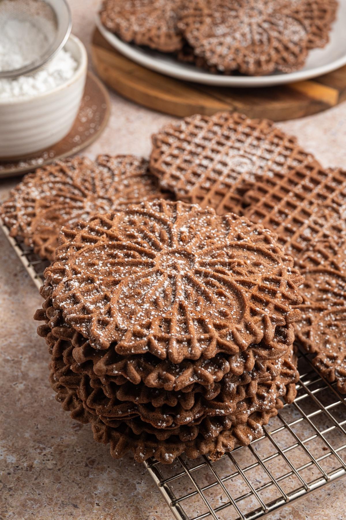 A stack of chocolate pizzelle cookies dusted with powdered sugar on a cooling rack.