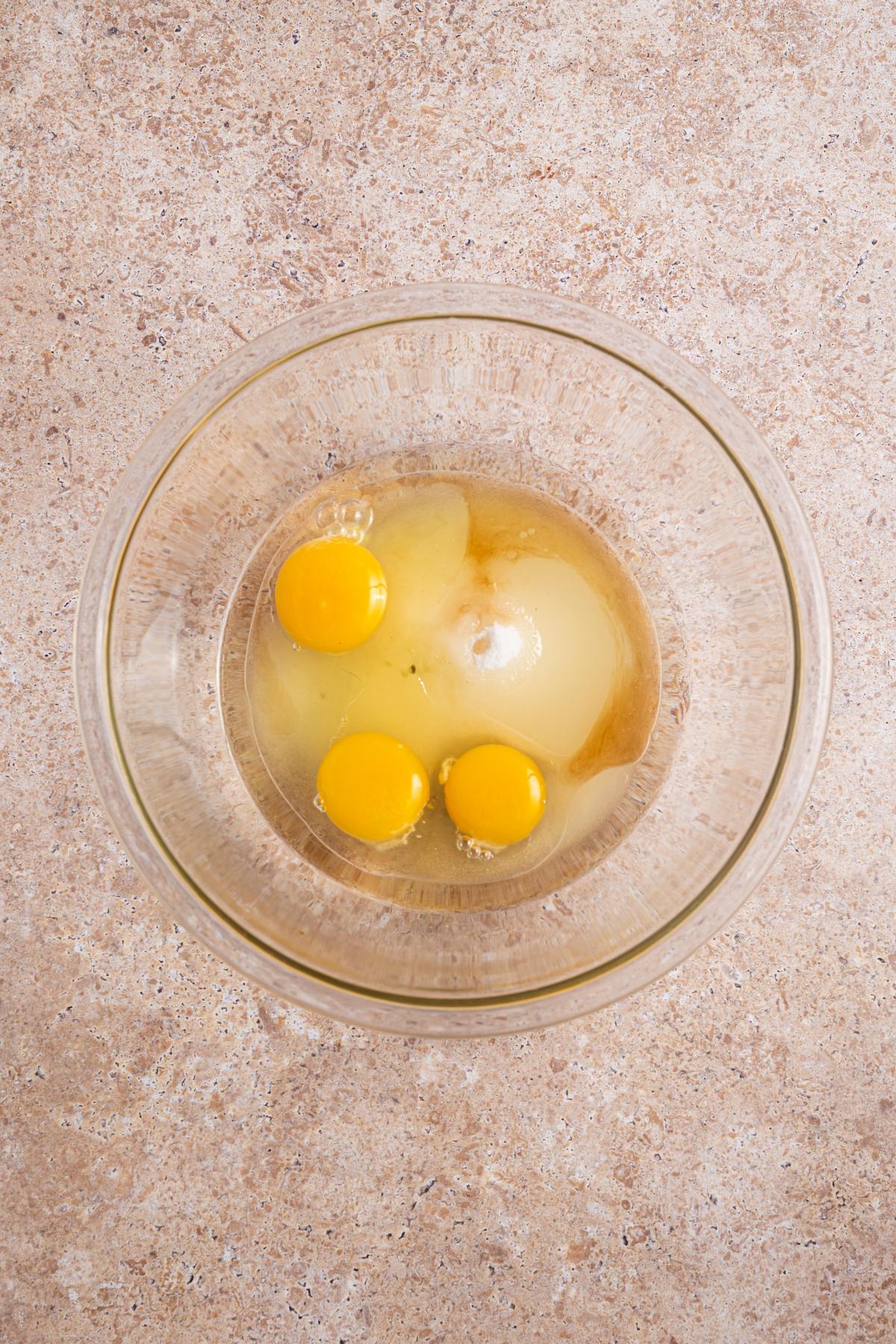 Three raw eggs in a glass bowl on a speckled beige countertop, viewed from above.