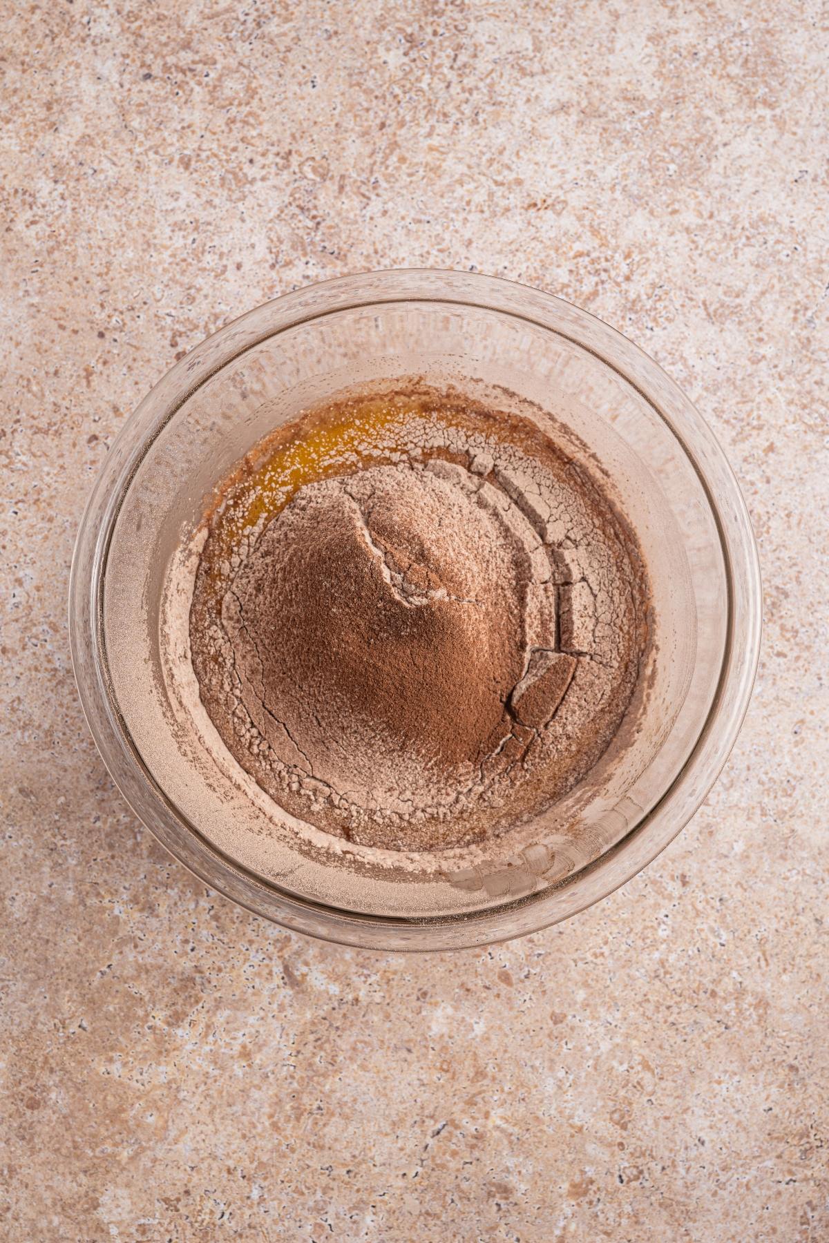 A glass bowl with cocoa powder and flour mixture on a beige countertop, viewed from above.