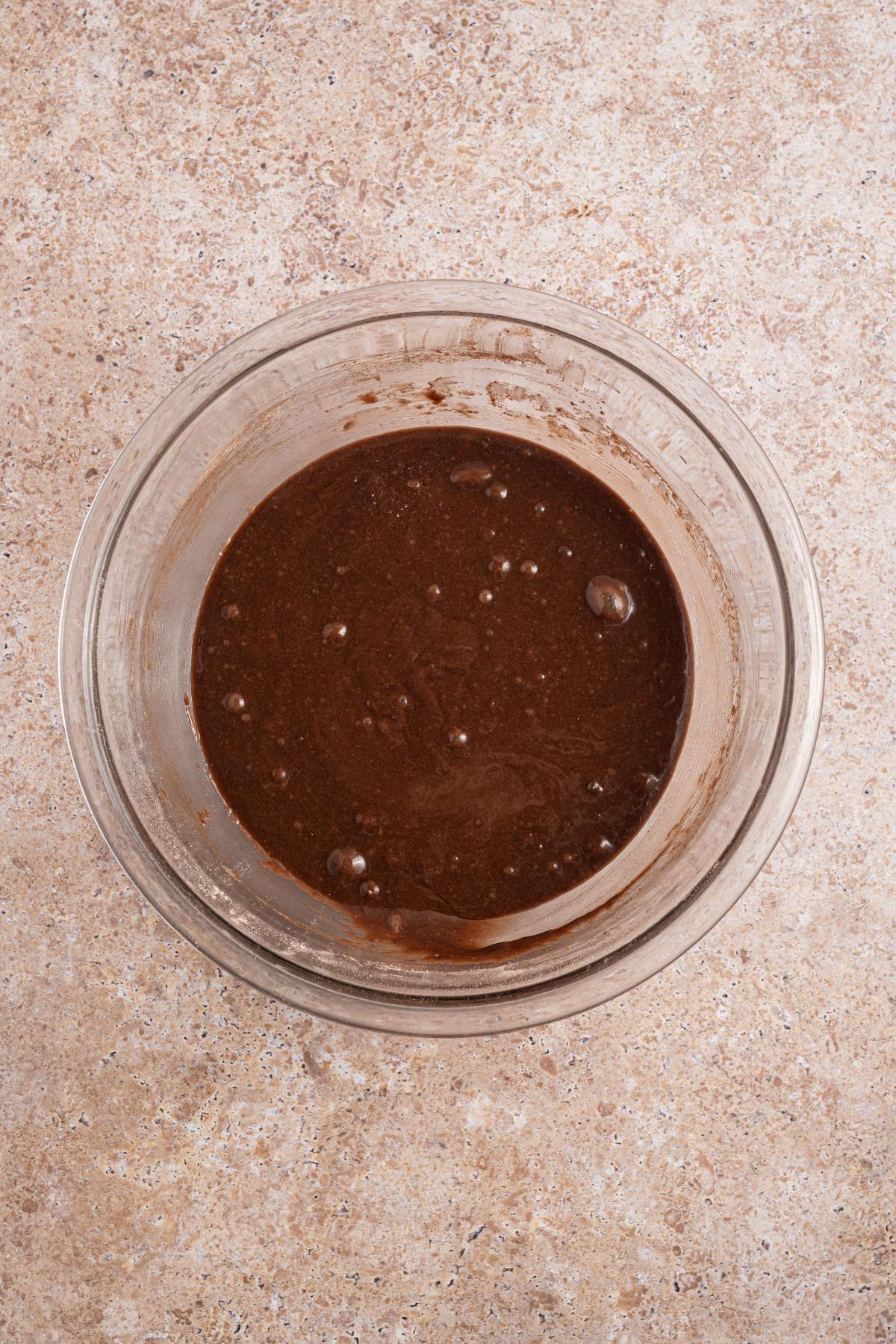 A glass bowl filled with chocolate cake batter on a light brown textured surface.