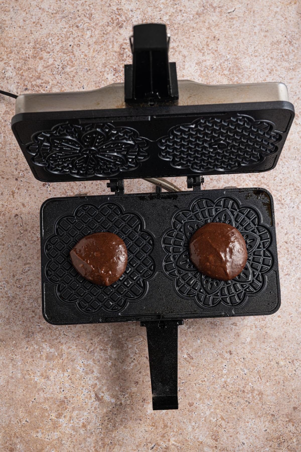 Open waffle iron with two scoops of chocolate batter on patterned plates, ready to be cooked.