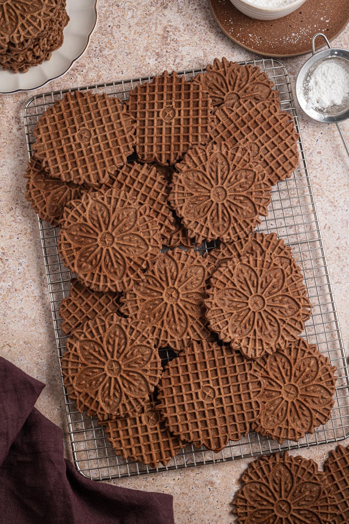 Chocolate pizzelle cookies with floral patterns on a cooling rack, surrounded by baking tools and a dark napkin.