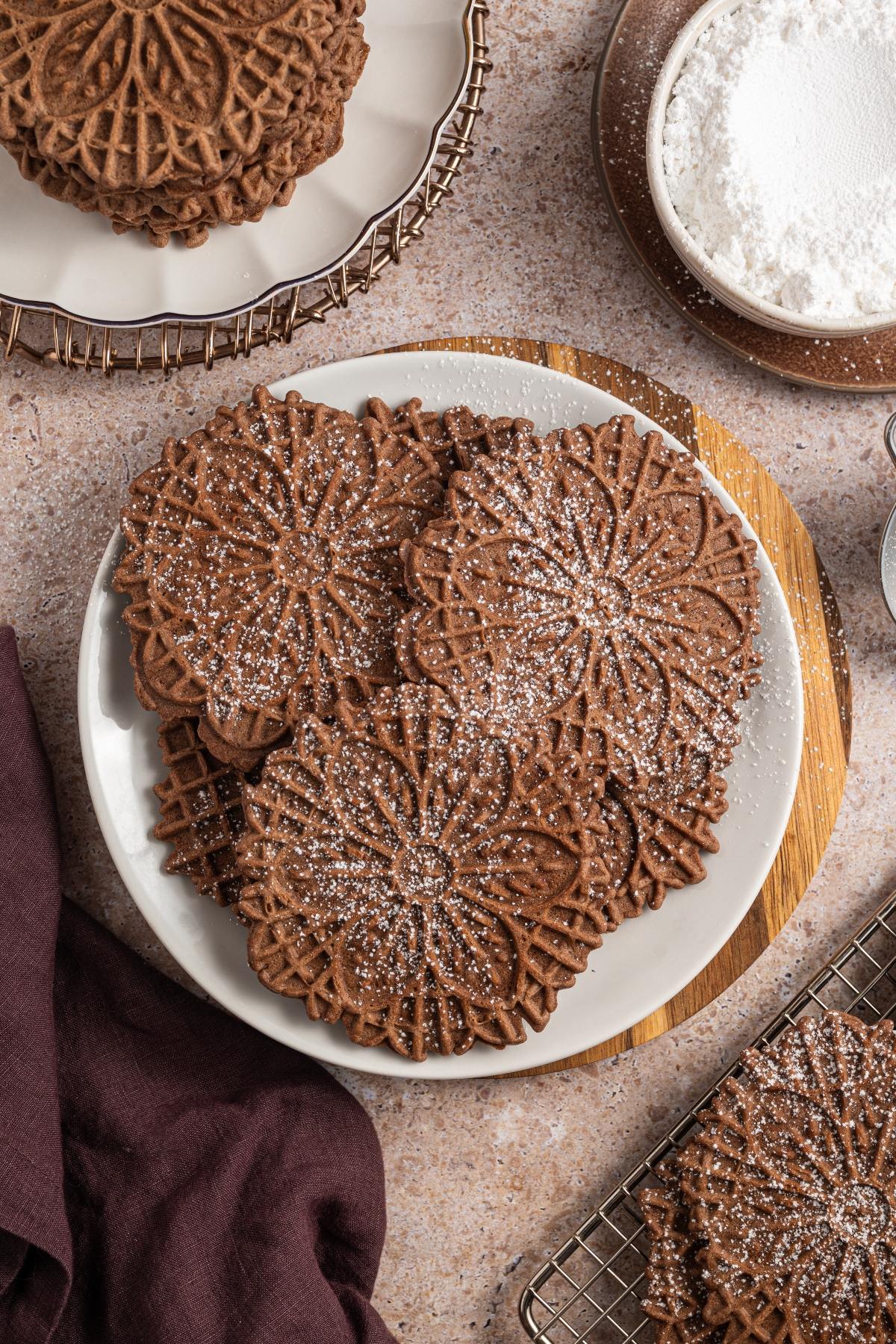 Chocolate pizzelle cookies dusted with powdered sugar are arranged on a white plate and a cooling rack.