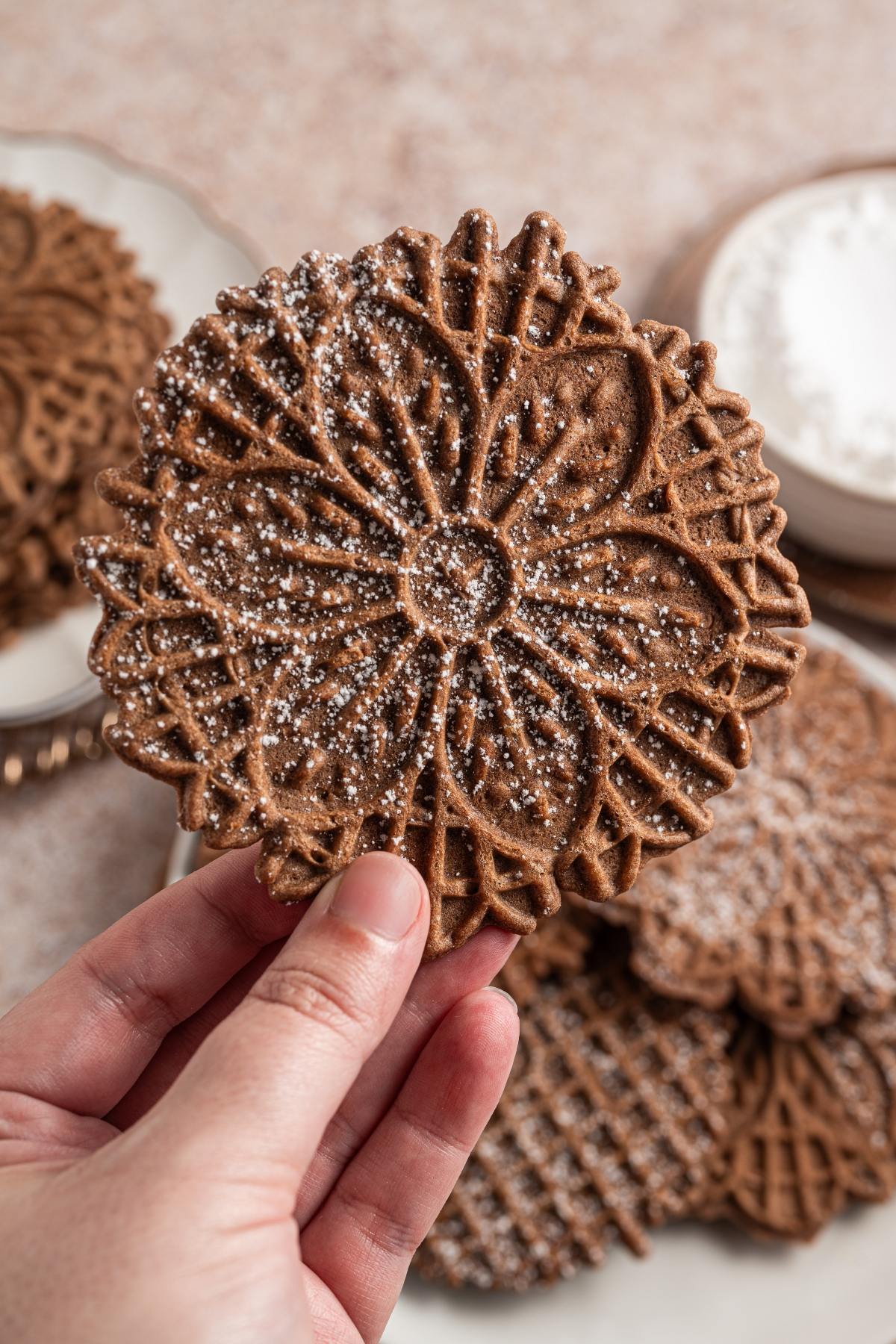 A hand holds a chocolate pizzelle cookie dusted with powdered sugar over a plate of more cookies.