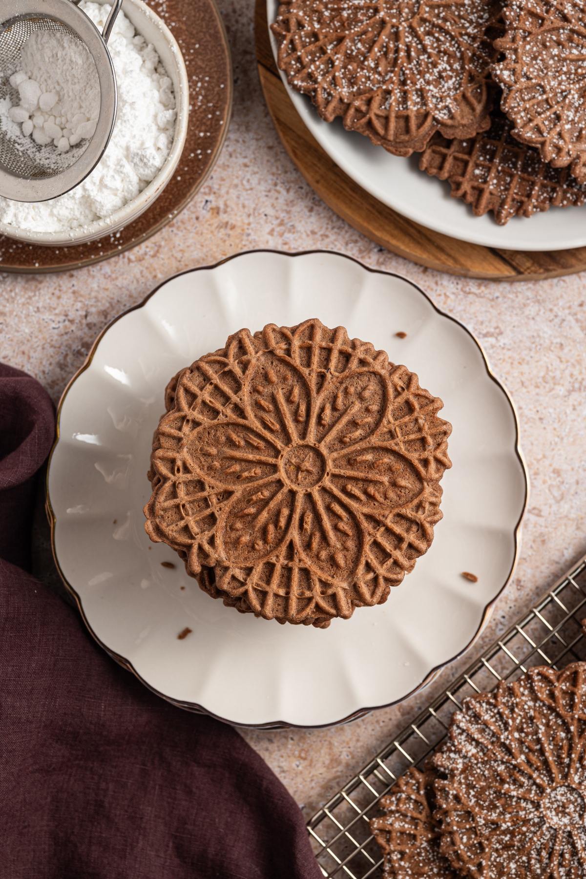 A stack of chocolate pizzelle cookies on a white plate, with powdered sugar and more cookies nearby.