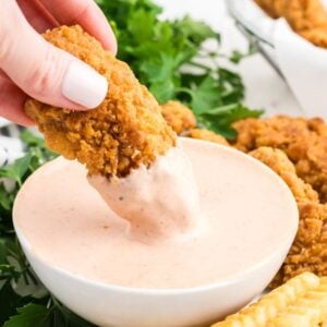 A hand dips a crispy chicken tender into a bowl of creamy Comeback Sauce, with golden fries and herbs nearby.