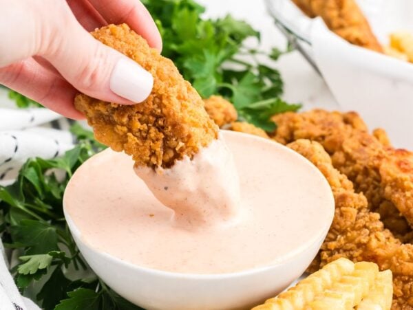 A hand dips a crispy chicken tender into a bowl of creamy Comeback Sauce, with golden fries and herbs nearby.