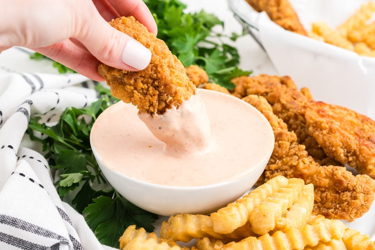Hand dipping a crispy chicken tender into a bowl of creamy Comeback Sauce, with crinkle-cut fries and parsley nearby.