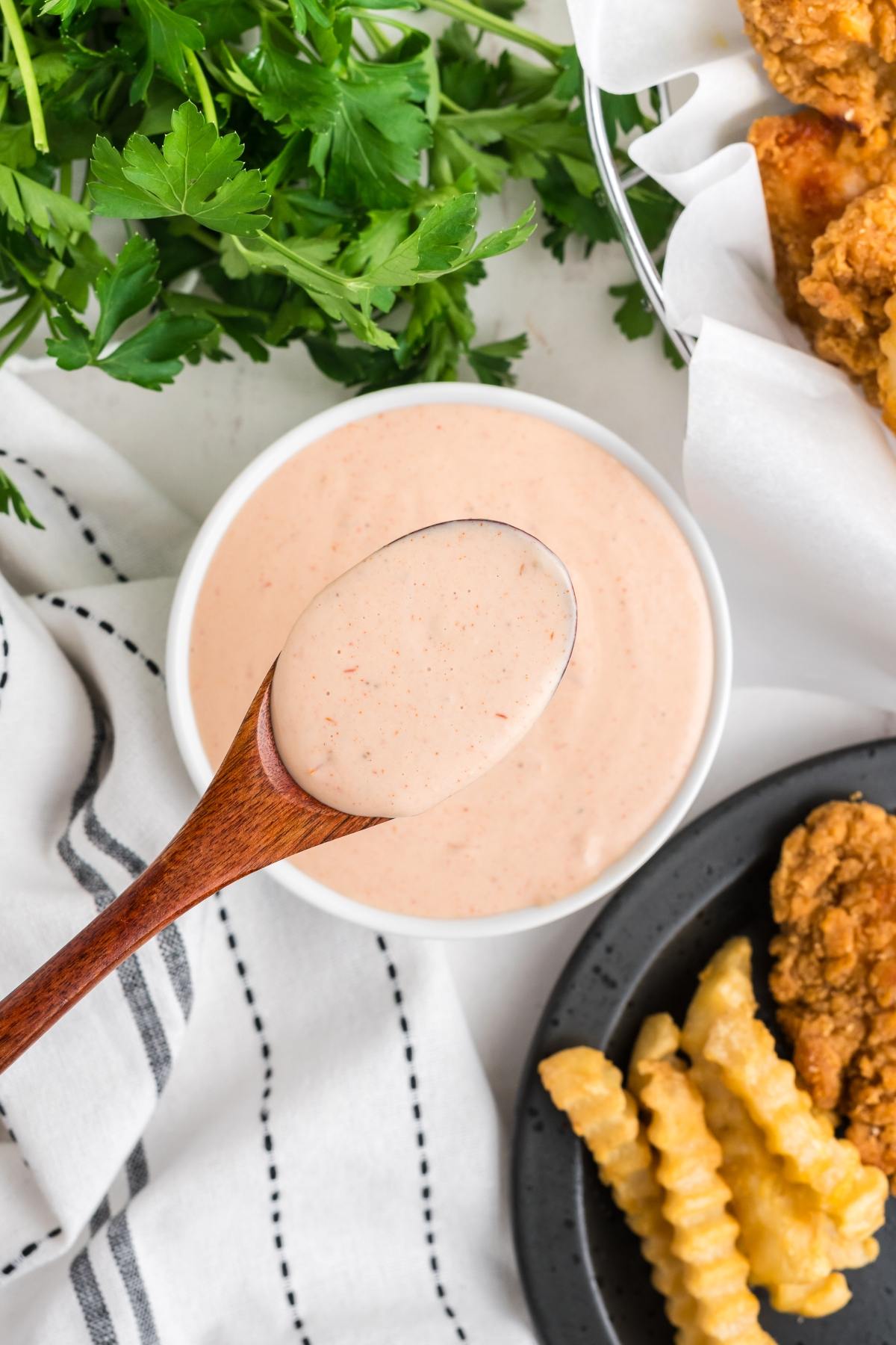 A spoon holds creamy dipping sauce above a bowl, with fries, fried chicken, and parsley nearby.