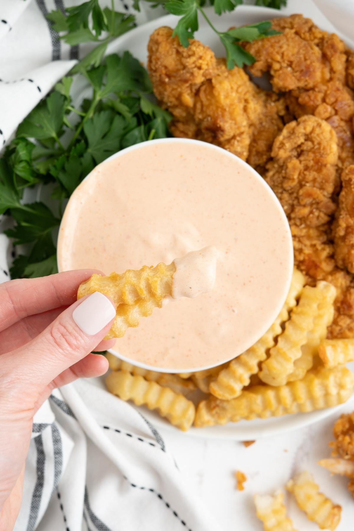 A hand dips a crinkle-cut fry into creamy sauce, with fried chicken and fries on a plate nearby.