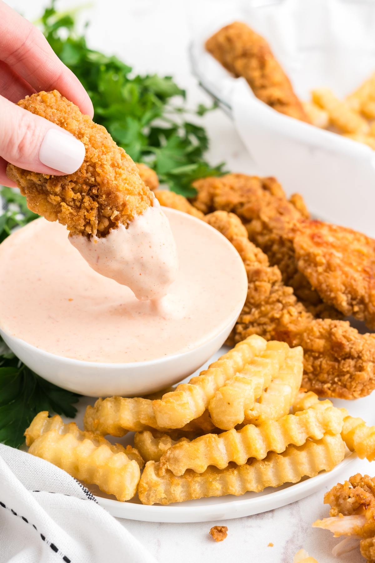 Hand dipping crispy fried chicken tender into creamy sauce, with crinkle-cut fries and more tenders nearby.