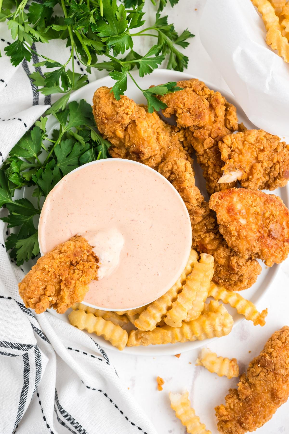 Plate with fried chicken tenders, crinkle-cut fries, parsley, and a bowl of dipping sauce with a tender in it.