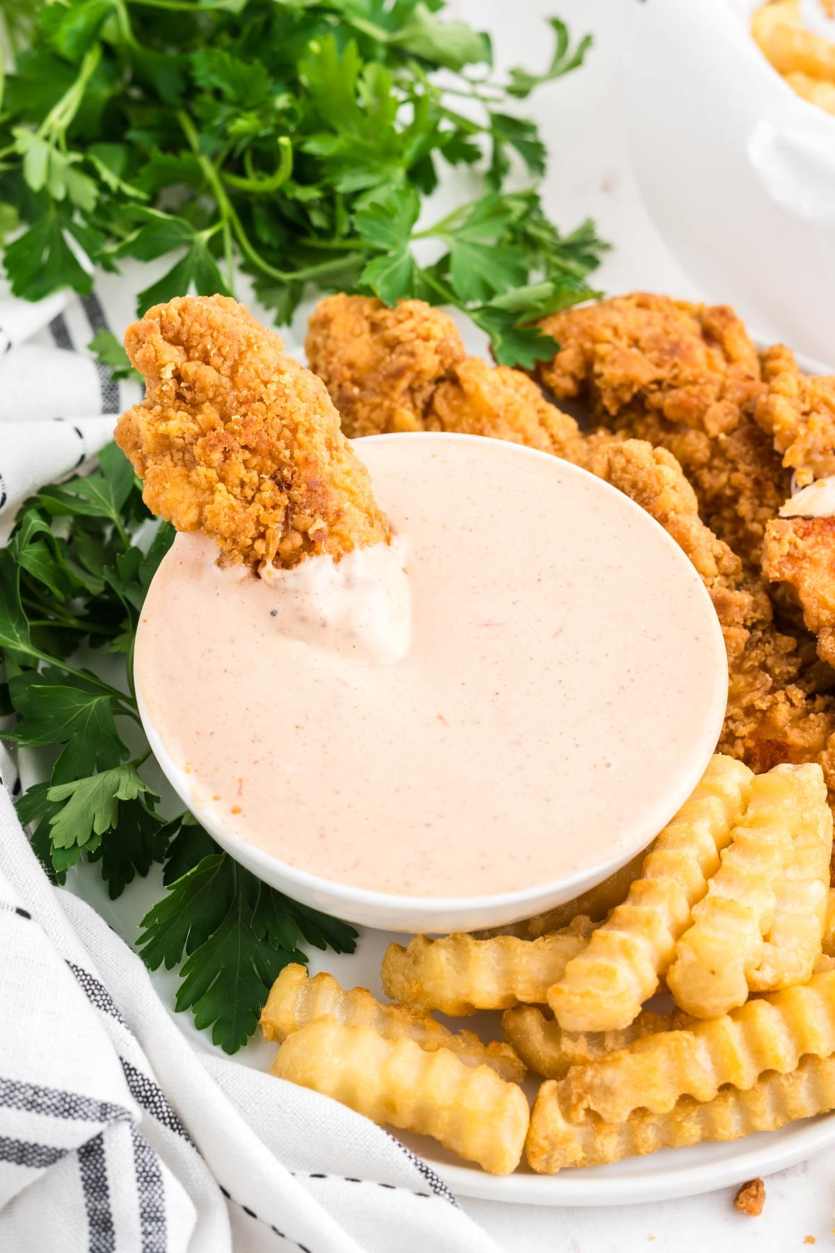 A chicken tender dipped in a creamy sauce, served with crinkle-cut fries and parsley on the side.