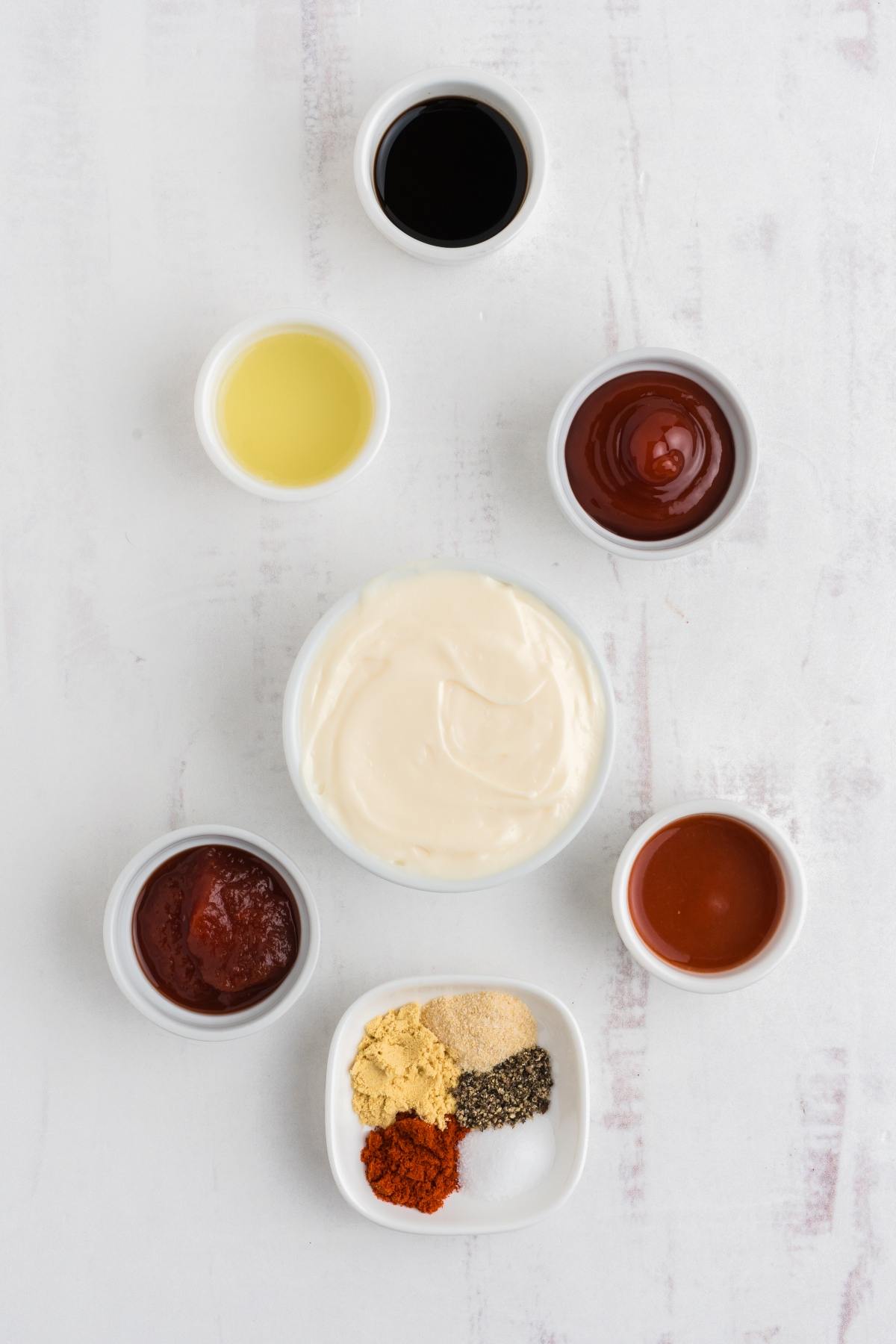 Seven small bowls with mayonnaise, ketchup, spices, mustard, barbecue sauce, hot sauce, and vinegar on a white surface.