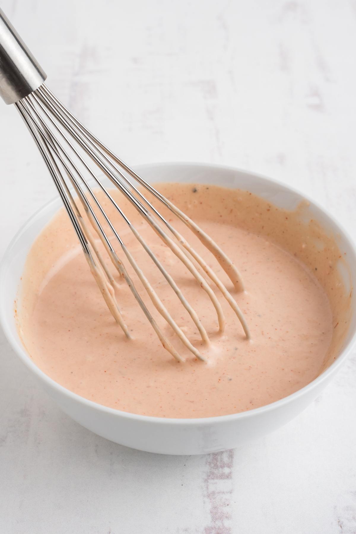 A metal whisk in a white bowl filled with creamy, light pink sauce on a white surface.