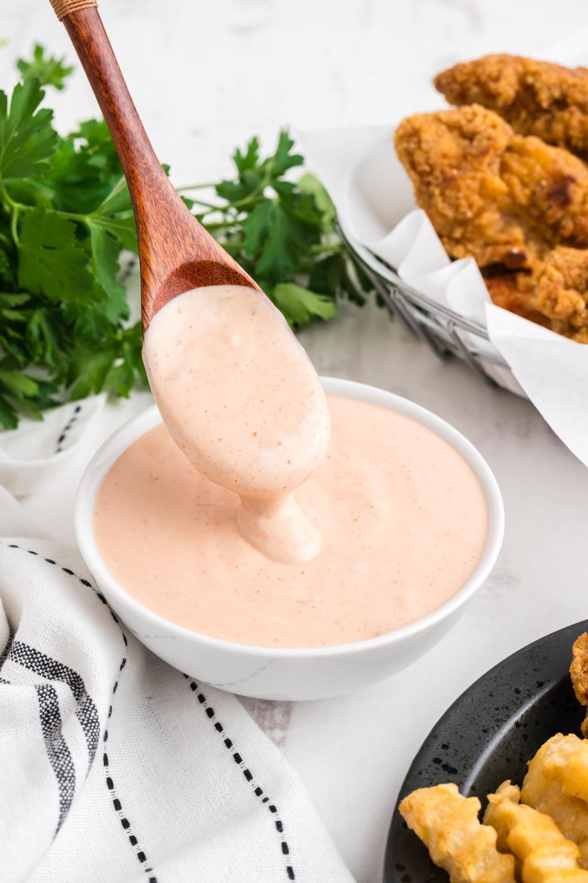 A wooden spoon scoops creamy dipping sauce from a bowl, with fried chicken and fries nearby.