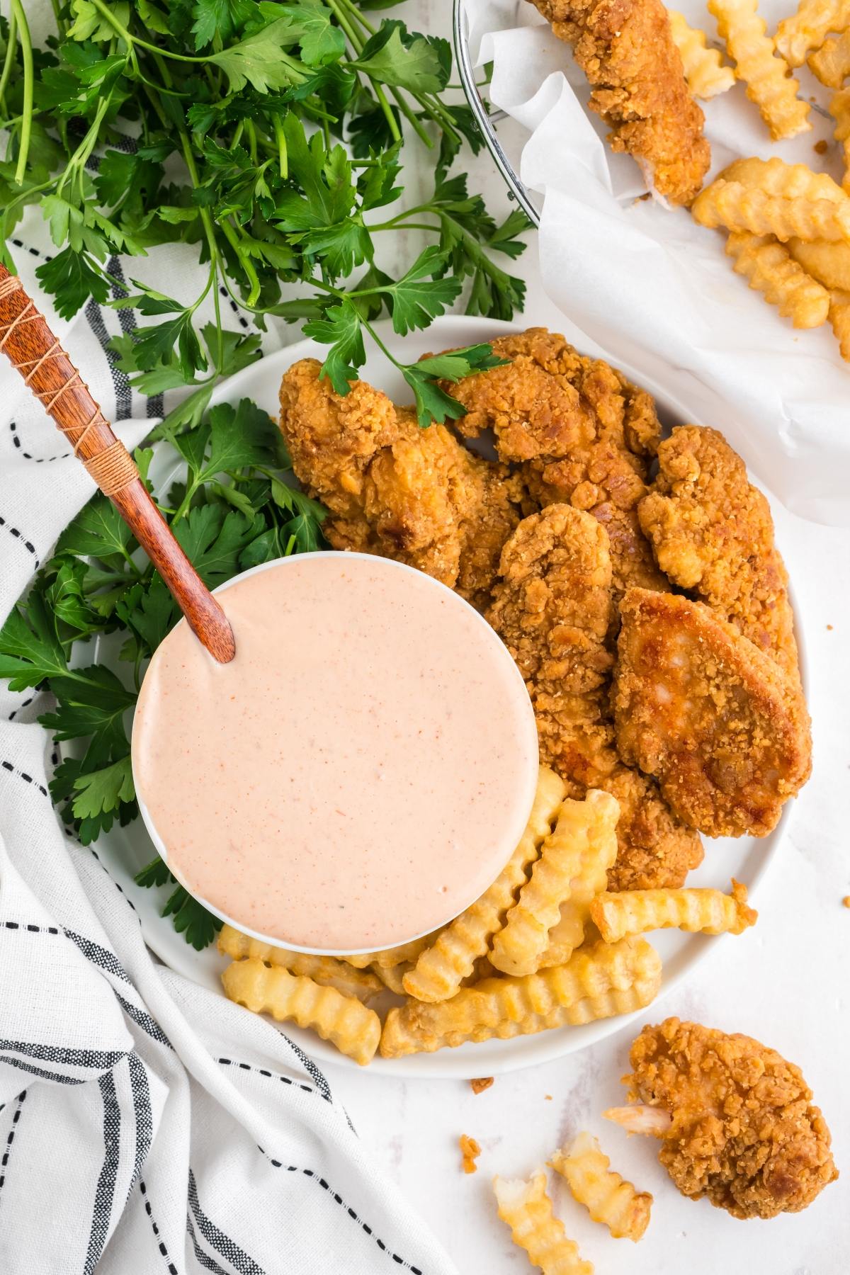 Plate of fried chicken, crinkle-cut fries, dipping sauce, and fresh parsley on a white surface.