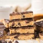 Stack of chocolate-topped Cookie Dough Bars with chocolate chips, surrounded by more bars and scattered chocolate chips.
