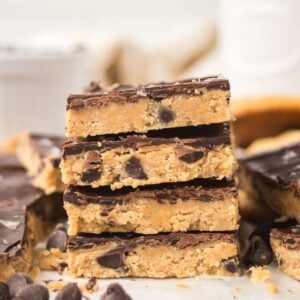Stack of chocolate-topped Cookie Dough Bars with chocolate chips, surrounded by more bars and scattered chocolate chips.